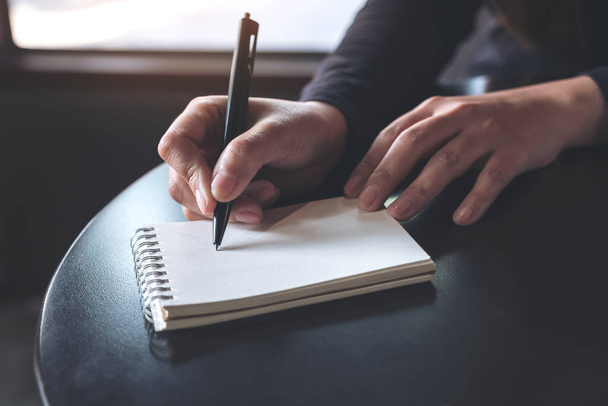 Szoros kép egy nő kezéről, amint egy fehér üres jegyzetfüzetre ír az asztalra.  - Fotó, kép