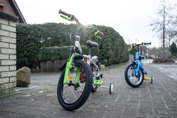 Two children's bicycles stand on the sidewalk in a small provincial town in Europe, in the background you can see the yard with a hedge and part of the house. - Photo, Image