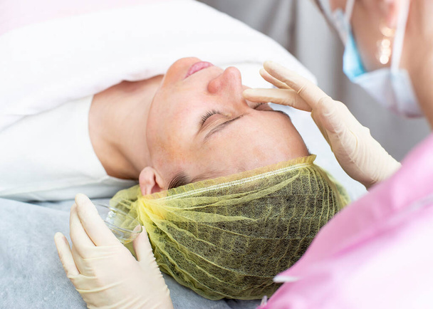 Een professionele cosmetoloog brengt een chemische peeling oplossing aan op de patiënt op de huid van het gezicht met behulp van de handen in handschoenen. Close-up. - Foto, afbeelding
