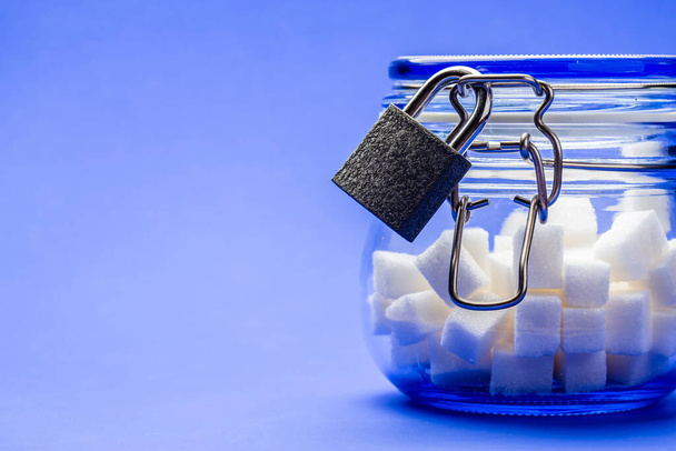 White sugar cubes in a transparent glass jar with a lid, which for restraint is closed on a metal padlock on a bright blue background with copy space. Concept of proper nutrition. Close-up. - Photo, Image
