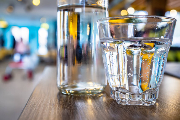Um copo e uma garrafa de água potável estão, para saciar a sede, em uma mesa de madeira, em um fundo embaçado brilhante. Close-up
. - Foto, Imagem