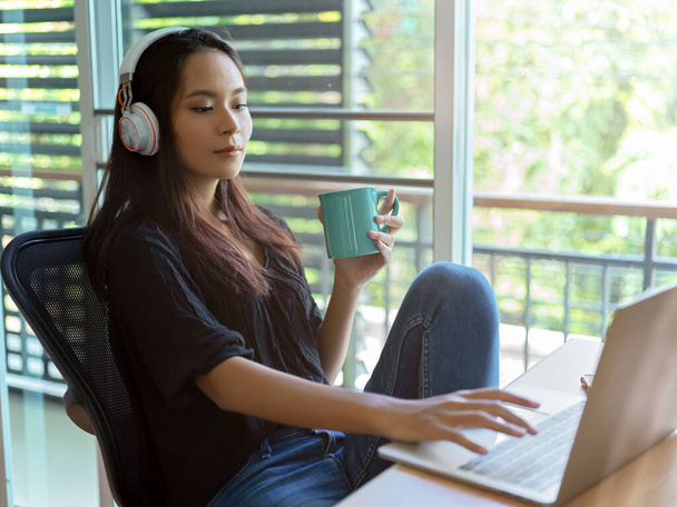 Side view of female relaxed woman with headphone using laptop and holding a coffee cup while sitting at worktable in bed room - Photo, Image