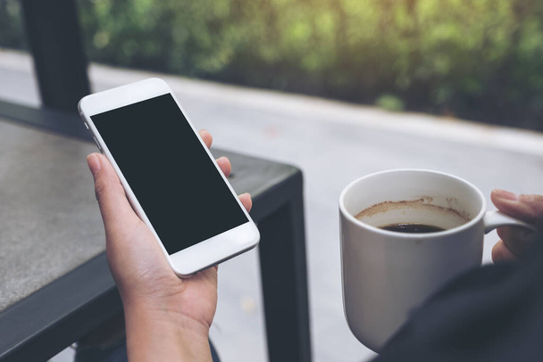 Imagen burlona de manos sosteniendo teléfono móvil blanco con pantalla de escritorio negro en blanco mientras bebe café en la cafetería
 - Foto, Imagen