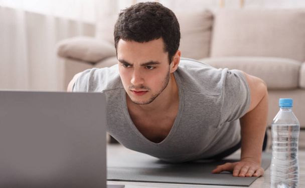 Moderní domácí cvičení s online lekcí. Mladý muž dělá kliky na podložce před notebookem, blízko vody - Fotografie, Obrázek
