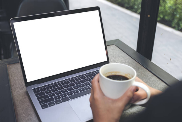 Mockup image of a woman using laptop with blank white desktop screen while drinking hot coffee in cafe - Photo, Image
