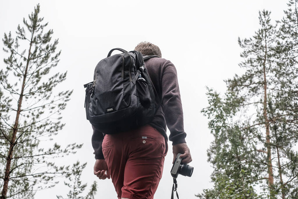 Чоловік-турист з рюкзаком і фотоапаратом в руці стоїть на вершині пагорба, на тлі похмурого неба і сосен. Крупним планом
. - Фото, зображення