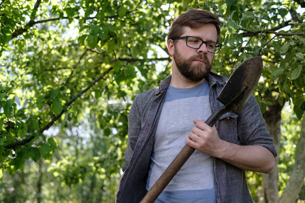 Egy töprengő ember kertész egy ásót vizsgál, amit a kezében tart, az almafák hátterében, egy nyári napon. Közelkép. - Fotó, kép