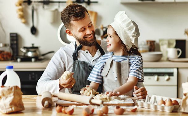 Glücklicher junger Mann mit kleiner Tochter in Kochmütze knetet Teig, während er Teig zubereitet und die gemeinsame Zeit in der heimischen Küche genießt - Foto, Bild
