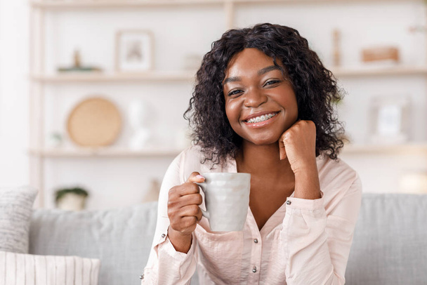 Radosna afrykańska kobieta relaksująca się na kanapie w domu z filiżanką kawy - Zdjęcie, obraz