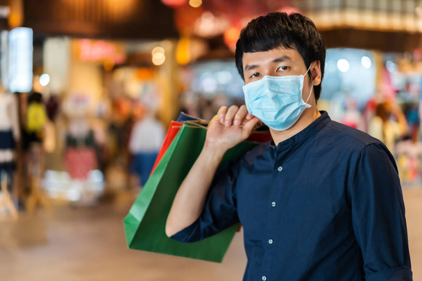 young asian man shopping with bag at mall and his wearing medical mask for prevention from coronavirus (Covid-19) pandemic. new normal concepts - Photo, image