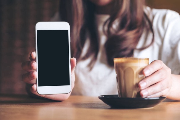 Mockup-Bild einer Frau, die weißes Handy mit leerem schwarzen Desktop-Bildschirm mit Kaffeetasse auf Holztisch hält und zeigt - Foto, Bild
