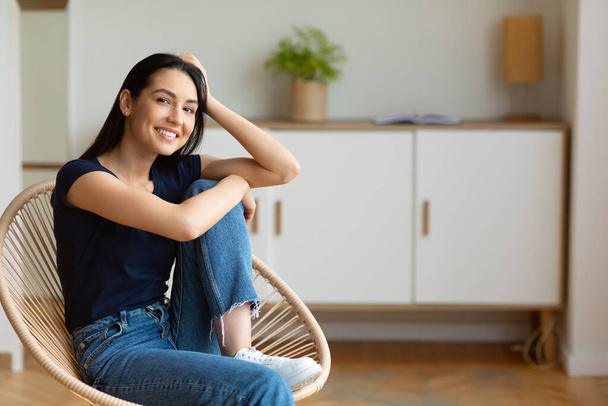 Chica sentada en silla moderna sonriendo a la cámara en casa
 - Foto, imagen