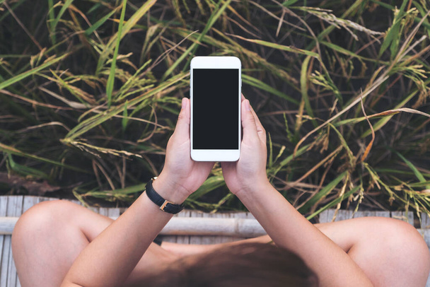 Вид зверху макет зображення рук жінки, що тримає білий мобільний телефон з порожнім чорним екраном робочого столу з зеленим фоном природи
 - Фото, зображення