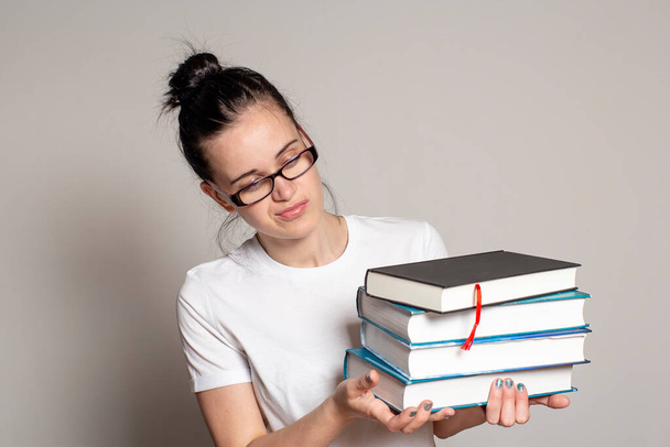 Piękna studentka w okularach trzyma stertę ciężkich książek w rękach i patrzy na nie zdziwiona. Izolacja na białym tle. - Zdjęcie, obraz