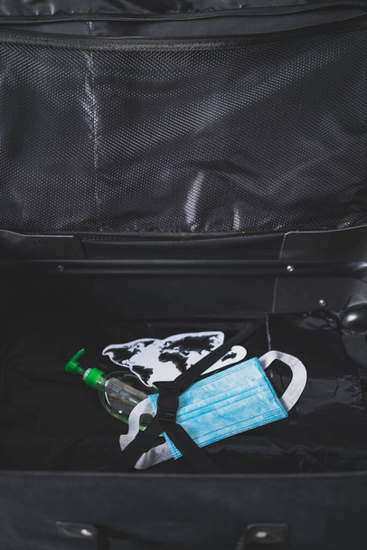 travel industry and flying during covid-19, carry-on luggage with world map next to surgical face mask and hand sanitizer for virus protection inside - Foto, imagen