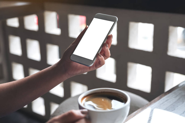 Mockup kuva naisen käsi tilalla valkoinen matkapuhelin tyhjä työpöytä näyttö ja kahvikuppi kahvilassa - Valokuva, kuva