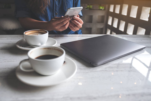 Imagen de cerca de una mujer sosteniendo y usando un teléfono inteligente con tazas de café y computadora portátil en la mesa en la cafetería
 - Foto, imagen