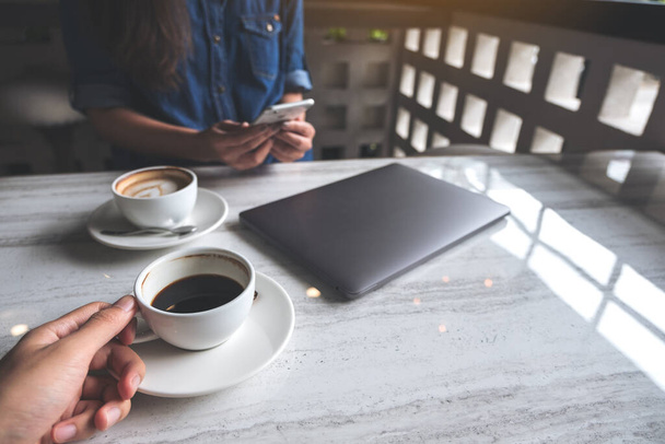 Крупный план мужчины, держащего чашку кофе, и женщины, использующей смартфон с ноутбуком на столе в кафе
 - Фото, изображение