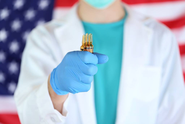 Coronavirusepidemie in de VS. Viral Disease Vaccine.Glass medische ampules in een hand in blauwe handschoenen op een Amerikaanse vlag wazig achtergrond - Foto, afbeelding