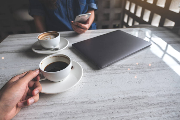 Szoros kép egy férfi kezében kávéscsésze és egy nő használ okostelefon laptop az asztalon kávézóban - Fotó, kép