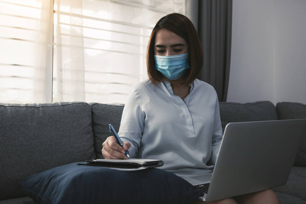 Azjatka pracująca w domu przy laptopie podczas epidemii koronawirusowej przez kwarantannę i nosząca maski medyczne. - Zdjęcie, obraz