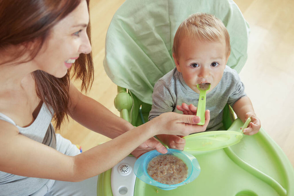 Glimlachende babysitter feed boy in kinderstoel met voedsel - Foto, afbeelding