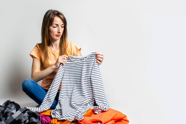 Jonge vrouw in T-shirt en jeans kiest dingen terwijl ze op de vloer zit op een lichte achtergrond. Verspreide dingen, keuze van kleding, keuze van stijl, geen geschikte kleding. - Foto, afbeelding