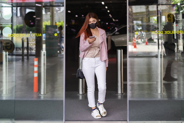 Красивые азиатские женщины носят черную маску. Используйте мобильный телефон ходить через стеклянные двери универмагов. - Фото, изображение