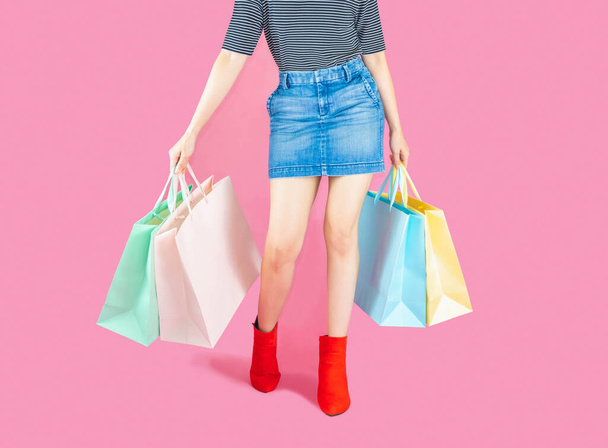 Sale and consumerism concept-Part body, beautiful female in denim short skirt, slender legs in red boots. Sexy woman holding a paper shopping bags, isolated on pink background - Φωτογραφία, εικόνα