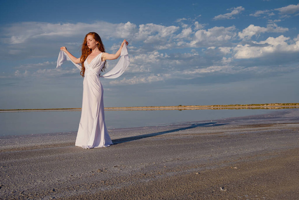 Beautiful young lady with long healthy red hair and cute white dress walking near lake. Hot summer evening. Sunset. Lifestyle concept. - Zdjęcie, obraz