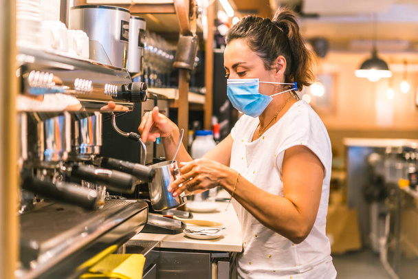 A waitress with a face mask in a bar. New normality, security measures after the coronavirus pandemic, social distance, covid-19. Making a coffee with milk - Foto, Imagem