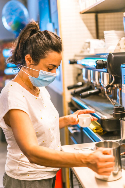Photo session with a waitress with a face mask in a bar. New normality, security measures after the coronavirus pandemic, social distance, covid-19. Preparing a coffee cut - Foto, afbeelding