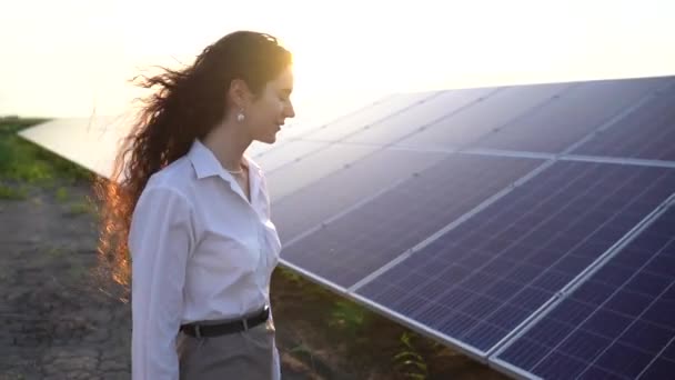 Gün batımında kadın güneş panellerinin yanında yürür ve güneş pillerine dokunur. Kadın yatırımcı resmi beyaz gömlek giyer. Ev için bedava elektrik. Gezegenin sürdürülebilirliği. Yeşil enerji. - Video, Çekim