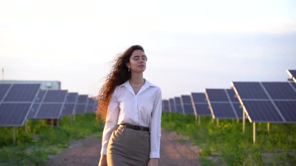 Tyttö kävellä välillä 2 Aurinkopaneelit rivi maassa auringonlaskun aikaan. Naissijoittaja käyttää virallista valkoista paitaa. Ilmainen sähkö kotiin. Planeetan kestävyys. Vihreä energia. - Materiaali, video