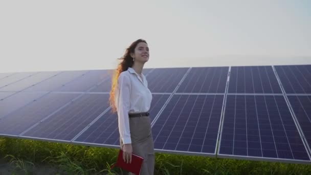 A nő napelemek közelében sétál a földön napnyugtakor, és mosolyog. A női befektető hivatalos fehér inget visel. Ingyen áram az otthonnak. A bolygó fenntarthatósága. Zöld energia. - Felvétel, videó
