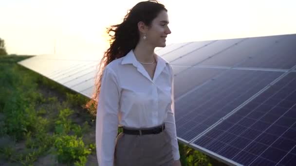 Nainen kävelee lähellä aurinkopaneelit rivi maassa auringonlaskun ja hymyillä ja katsoa aurinkokennoja. Naissijoittaja käyttää virallista valkoista paitaa. Ilmainen sähkö kotiin. Planeetan kestävyys. Vihreä energia. - Materiaali, video
