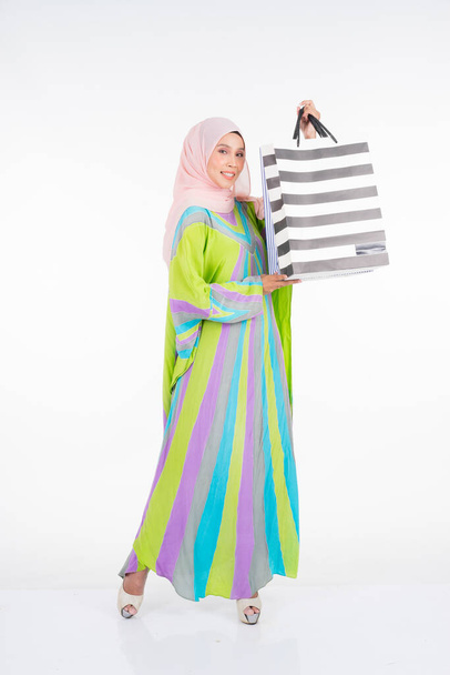Ένα όμορφο και χαρούμενο μουσουλμανικό γυναικείο μοντέλο με παραδοσιακό καφτάνι / καφτάνι φόρεμα που μεταφέρει τσάντες για ψώνια απομονωμένες σε λευκό φόντο. Eidul fitri μόδα και εορταστική προετοιμασία ψώνια έννοια - Φωτογραφία, εικόνα