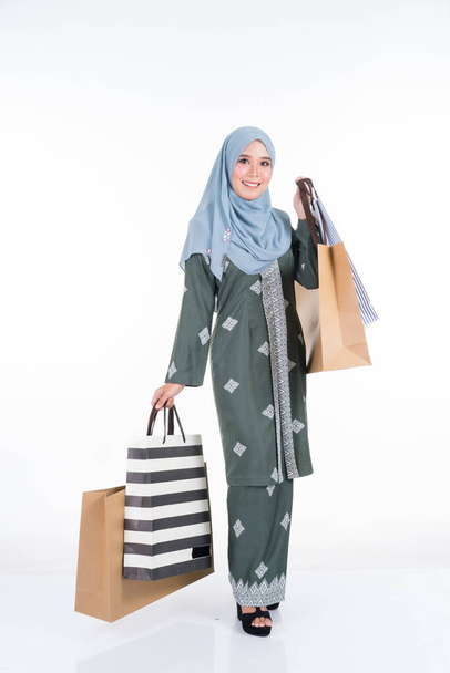 A beautiful and excited Muslim female model in a Asian traditional dress modern kebaya carrying shopping bags isolated on white background. Eidul fitri fashion and festive preparation shopping concept - Foto, immagini