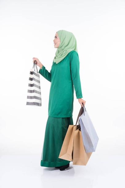 A beautiful Asian female model in a modern kurung carrying shopping bags isolated on white background. Eidul fitri festive preparation shopping concept. Full length portrait. - Fotoğraf, Görsel