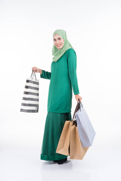A beautiful Asian female model in a modern kurung carrying shopping bags isolated on white background. Eidul fitri festive preparation shopping concept. Full length portrait. - Foto, Bild
