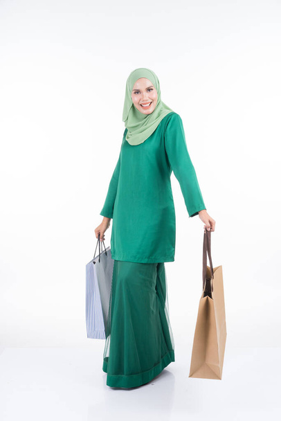 A beautiful Asian female model in a modern kurung carrying shopping bags isolated on white background. Eidul fitri festive preparation shopping concept. Full length portrait. - Fotografie, Obrázek