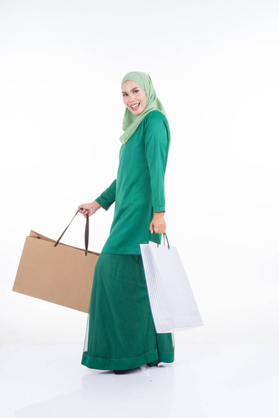 A beautiful Asian female model in a modern kurung carrying shopping bags isolated on white background. Eidul fitri festive preparation shopping concept. Full length portrait. - Photo, Image