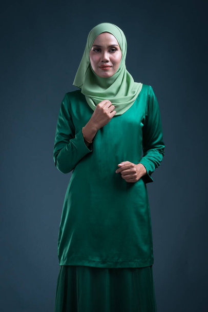 Halve lengte portret van een prachtig vrouwelijk moslim model in moderne kurung en hijab, een urban lifestyle kleding voor moslimvrouwen op groene achtergrond. Schoonheid en hijab mode portret concept. - Foto, afbeelding