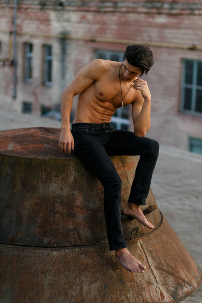 Handsome shirtless, sexy brunette young man with tanned athletic body, seated in profile on old roof at the sunset. - Foto, Bild