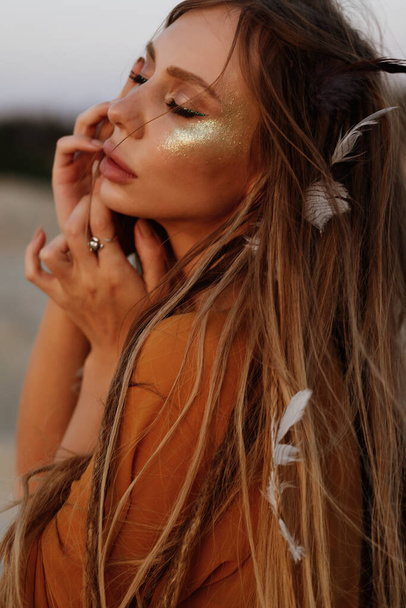 modelo bronzeado com dreadlocks e penas no cabelo com lantejoulas de ouro em seu rosto
 - Foto, Imagem