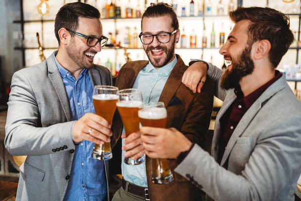 Happy young successful businessmen drinking beer and talking at pub after work - 写真・画像