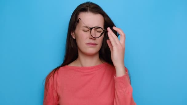 Tired of eyeglasses young woman massaging nose bridge, exhausted lady suffering from headache or migraine, feeling eye strain, wears red sweater, isolated on blue background. Eyes fatigue concept - Filmati, video