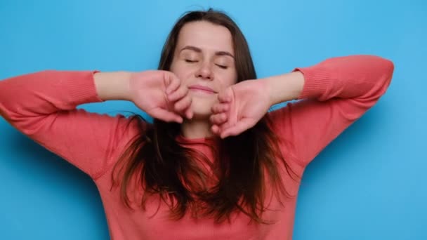 Cute young woman smiles broadly, has sensual look, keeps hand near cheek, eyes closed, romantic expression, remembers something touching or pleasant, wears yellow sweater, isolated on blue background - Footage, Video
