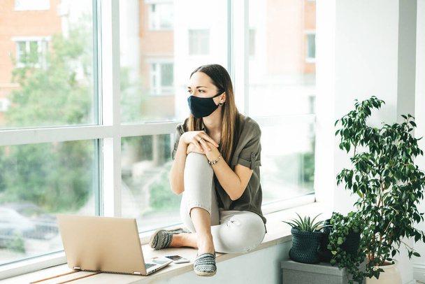 Droevig meisje werknemer of student zit aan het raam met een masker met een computer. Ze bleef thuis of werkt bij het raam in een open ruimte. - Foto, afbeelding