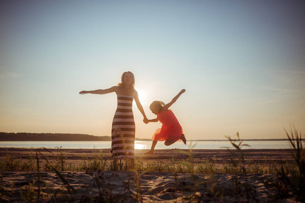 Joyeux maman et fille en robes sont sauter et danser sur la plage pendant le coucher du soleil. De bonnes relations depuis deux générations. Amusant passe-temps familial dans la nature. Promotion de la santé par des jeux et des activités de plein air. - Photo, image